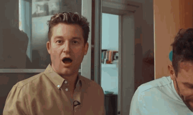 a man in a tan shirt with a microphone on his neck looks surprised