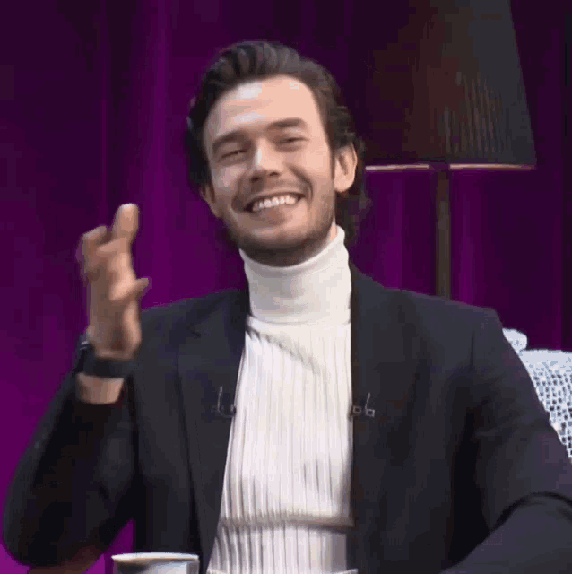 a man wearing a black jacket and a white turtleneck is sitting in front of a purple curtain