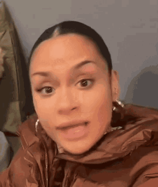 a woman in a brown jacket and earrings is making a funny face .