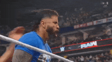a man in a blue shirt is standing in a wrestling ring with a crowd in the background .