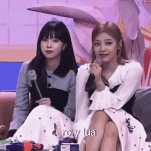 two women are sitting next to each other on a couch holding microphones and smiling .