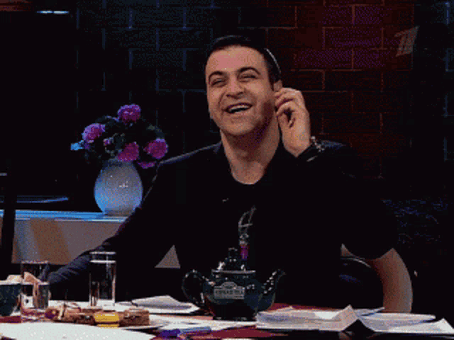 a man is sitting at a table talking on a cell phone with a vase of flowers in the background