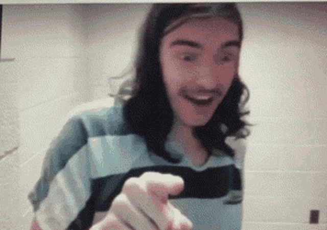 a man with long hair and a mustache is smiling and pointing at the camera
