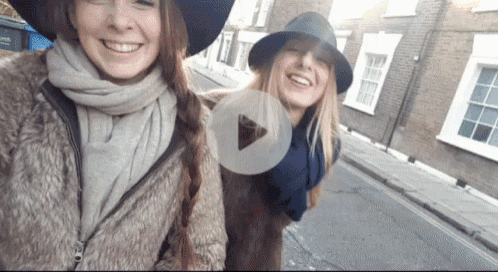 two women standing next to each other on a street with a play button in the corner