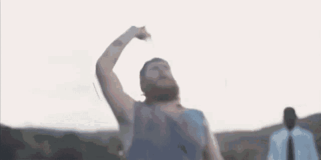 a man with a beard is standing in a field with his arms in the air and giving the middle finger .