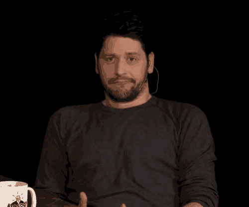 a man with a beard is making a funny face while sitting at a table with a coffee mug .