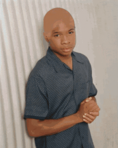 a bald man in a blue shirt stands with his hands folded