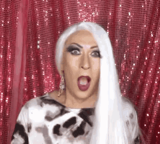a drag queen with white hair is making a surprised face in front of a pink curtain
