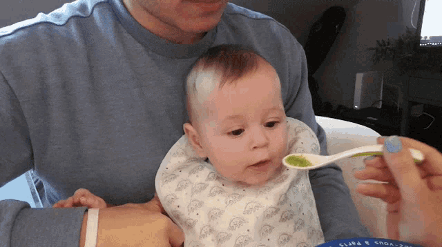 a baby in a bib is being fed with a spoon by a man in a blue shirt with the word paris on the sleeve