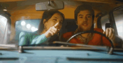a man and a woman are sitting in a car and the woman is pointing at the camera .