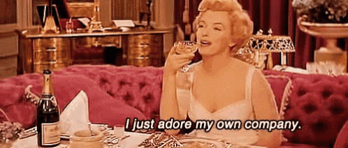 a woman is sitting at a table drinking a glass of wine and saying `` i just adore my own company '' .