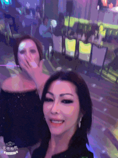 two women are posing for a selfie in a room with chinese writing on the bottom right