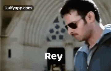 a man wearing sunglasses is standing in front of a building with the word rey written on the bottom .