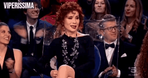 a woman in a black dress is sitting in front of a crowd of people applauding .