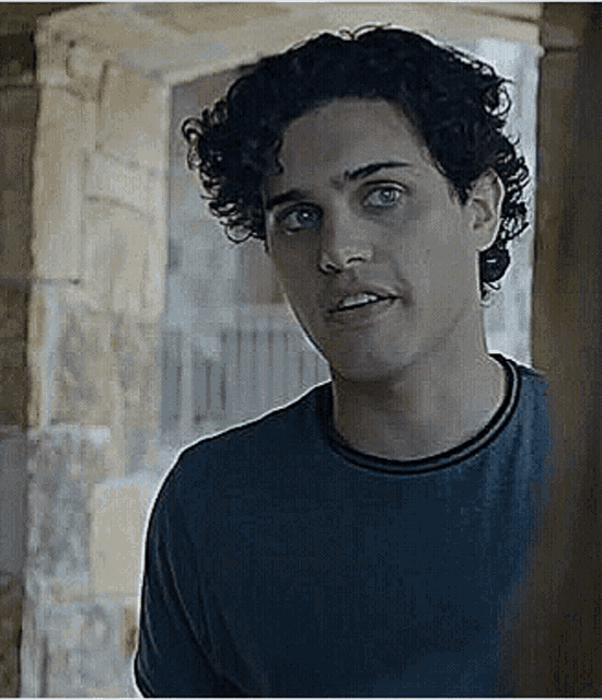 a young man with curly hair and blue eyes is standing in front of a window .