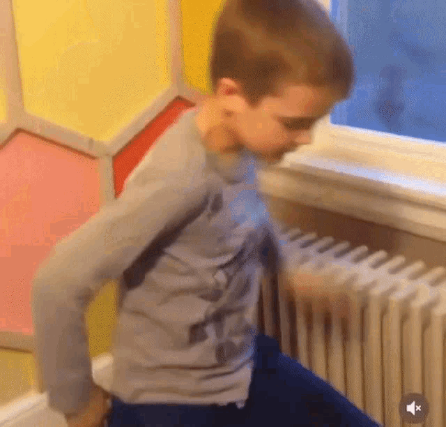 a young boy in a gray shirt is standing next to a radiator in front of a window .
