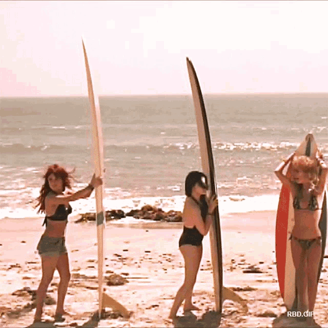 three women holding surfboards on a beach with rbd.gif in the bottom right corner
