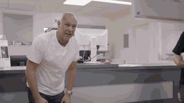 a bald man in a white shirt stands in front of a computer screen