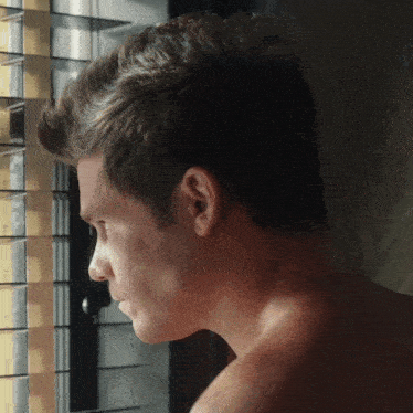 a man without a shirt is looking out a window with blinds