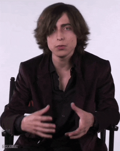 a young man with long hair is sitting in a chair with his hands folded .