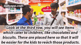 a man is talking to another man in a store while looking at items which cater to children like chocolates and biscuits .