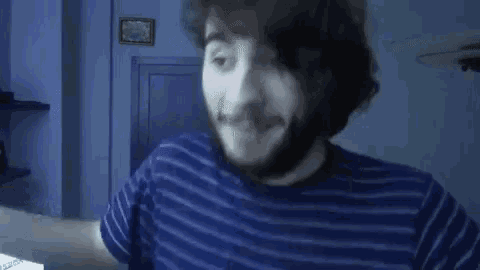 a man with a beard wearing a blue striped shirt is standing in front of a computer screen .