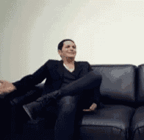 a man is sitting on a black leather couch with his legs crossed and smiling .