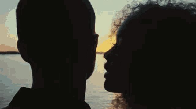 a man and a woman are looking at each other while standing next to a body of water at sunset .