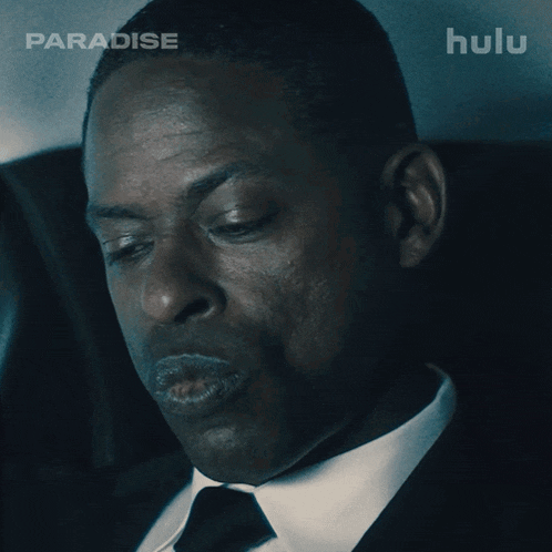 a man in a suit and tie is sitting in a car with hulu behind him