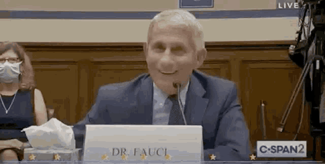a man in a suit and tie is sitting at a table with a sign that says `` dr. fauci '' .