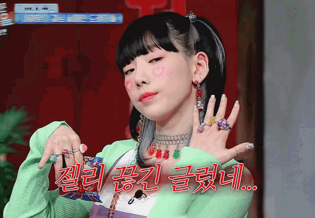 a woman wearing a green sweater shows off her rings and earrings with korean writing behind her