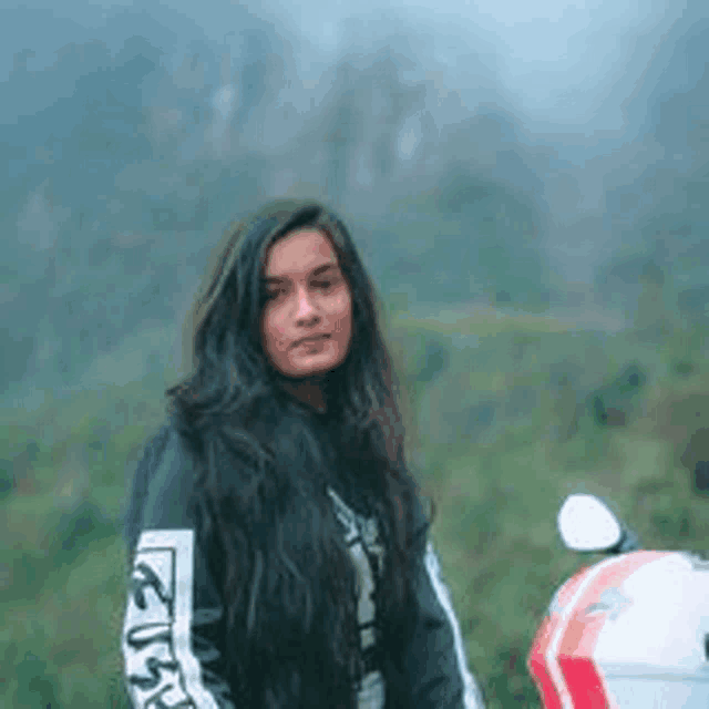 a woman with long hair is standing next to a motorcycle in the woods .