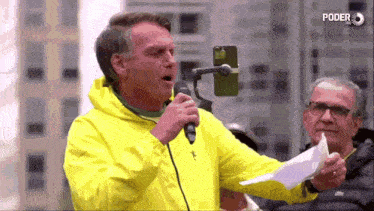 a man in a yellow jacket is talking into a microphone while holding a piece of paper .