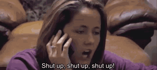 a woman is sitting on a couch talking on a cell phone and saying shut up .