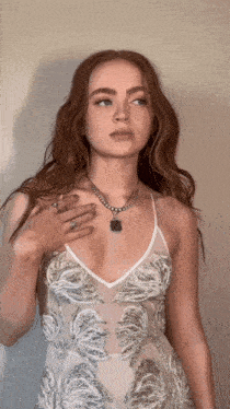 a woman wearing a white dress and a necklace holds her hand to her chest