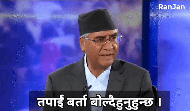 a man wearing glasses and a black hat is sitting in front of a sign that says ranjan on it