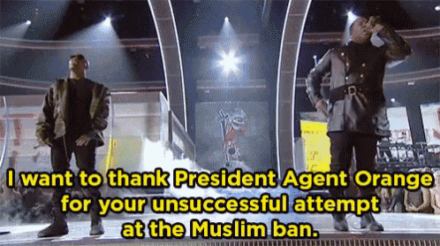 two men standing on a stage with the words " i want to thank president agent orange for your unsuccessful attempt at the muslim ban " above them