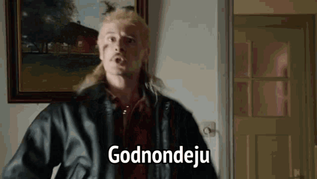 a man with a mullet and a leather jacket is standing in front of a painting and saying godnondjeju .