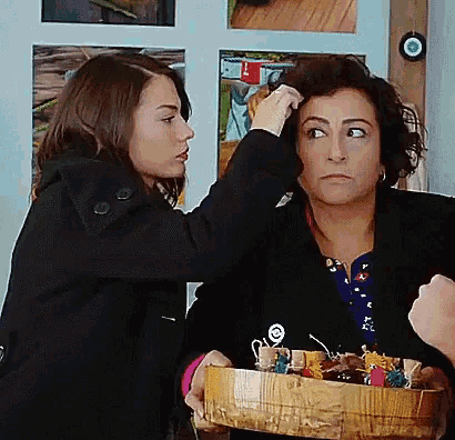 a woman in a black coat is touching another woman 's hair while holding a wooden box