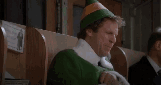 a man dressed as an elf is laughing while sitting in a chair next to another man in a suit .