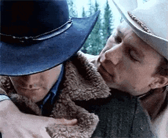 two men in cowboy hats are hugging each other . one of the men is wearing a blue hat .