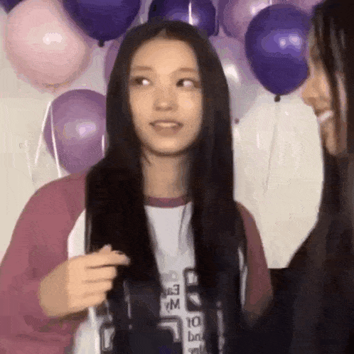 two girls are standing next to each other in front of balloons . one girl is holding another girl 's hair .