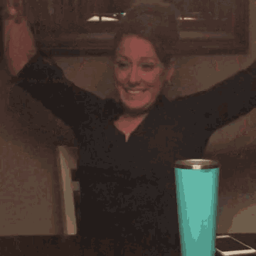 a woman is sitting at a table with her arms in the air and a blue cup in front of her .