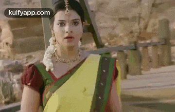a woman in a yellow and green saree is standing in front of a building and looking at the camera .
