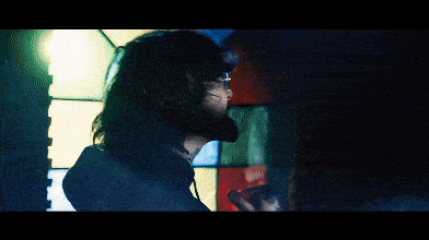 a man with long hair is standing in front of a stained glass window with the letter k on it