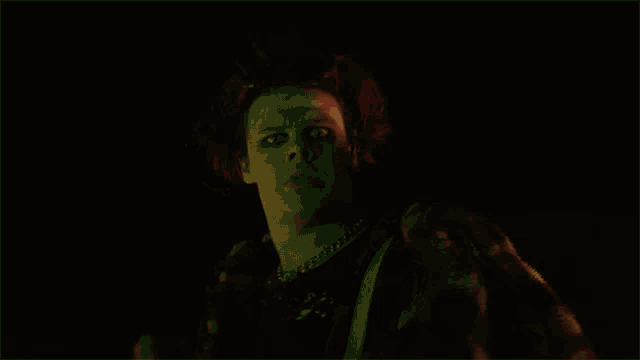 a young man with a chain around his neck looks at the camera in a dark room