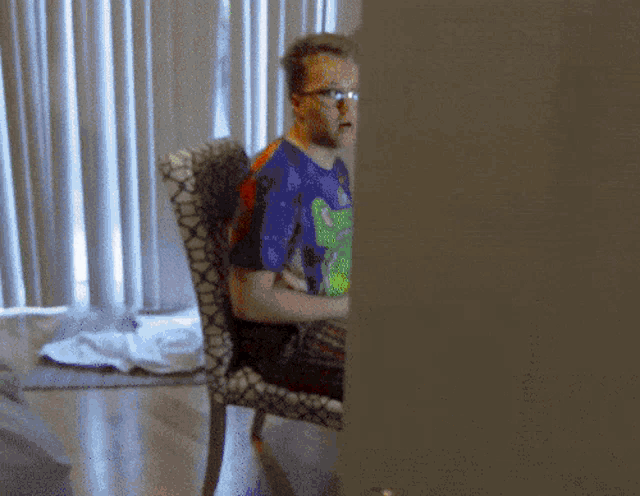 a man wearing glasses and a blue shirt with a frog on it is sitting in a chair