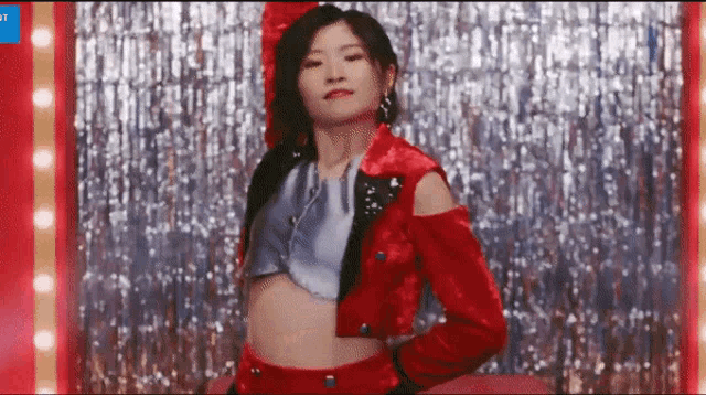 a woman in a red jacket and crop top is dancing in front of a curtain of lights .