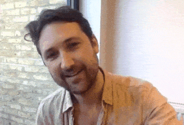 a man with a beard is smiling for the camera while sitting at a table in front of a window .