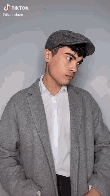 a young man wearing a gray coat and a hat is standing in front of a wall .